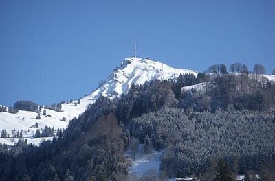 Verschneiter Berggipfel mit Sendemast über bewaldeten Hängen unter blauem Himmel.
