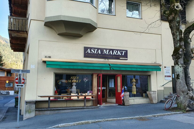 <p>Ein asiatischer Markt mit einem Schaufenster und einer rot lackierten Tür. Vor dem Geschäft stehen ein Fahrrad und einige Produkte sind ausgestellt.</p>