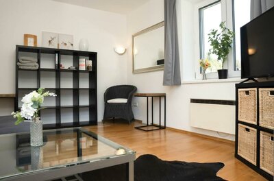 Ein modernes Wohnzimmer mit einem gläsernen Tisch, einem schwarzen Regal und einem Sofa. Große Fenster lassen viel Licht herein und sorgen für eine einladende Atmosphäre.
