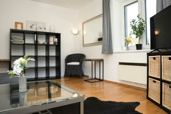 Ein modernes Wohnzimmer mit einem gläsernen Tisch, einem schwarzen Regal und einem Sofa. Große Fenster lassen viel Licht herein und sorgen für eine einladende Atmosphäre.