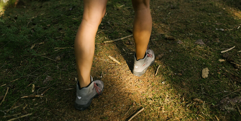 Eine Person mit grauen Wanderschuhen bewegt sich durch einen ruhigen Wald, der mit grünem Moos und kleinen Zweigen bedeckt ist.