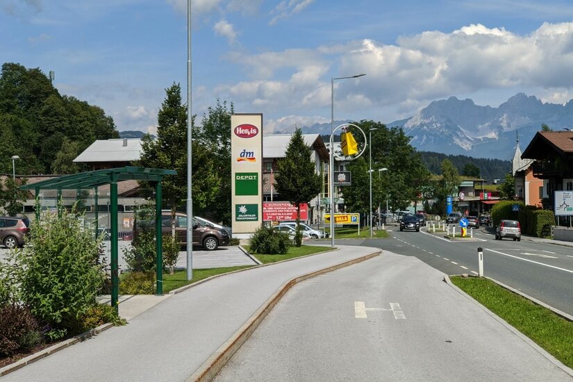 Blick auf eine Bushaltestelle und einen Gehweg vor einem Fachmarktzentrum mit Bergpanorama im Hintergrund.