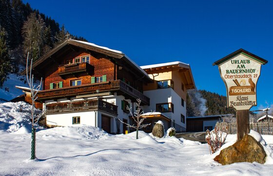 Ein traditionelles Bauernhaus aus Holz und Stein in einer verschneiten Winterlandschaft unter blauem Himmel.