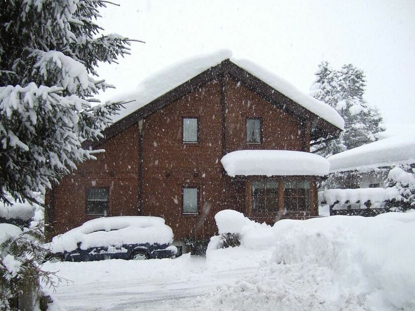 Haus Pirchner im Winter Ein braunes Holzhaus mit schneebedecktem Dach und ein eingeschneites Auto bei starkem Schneefall.