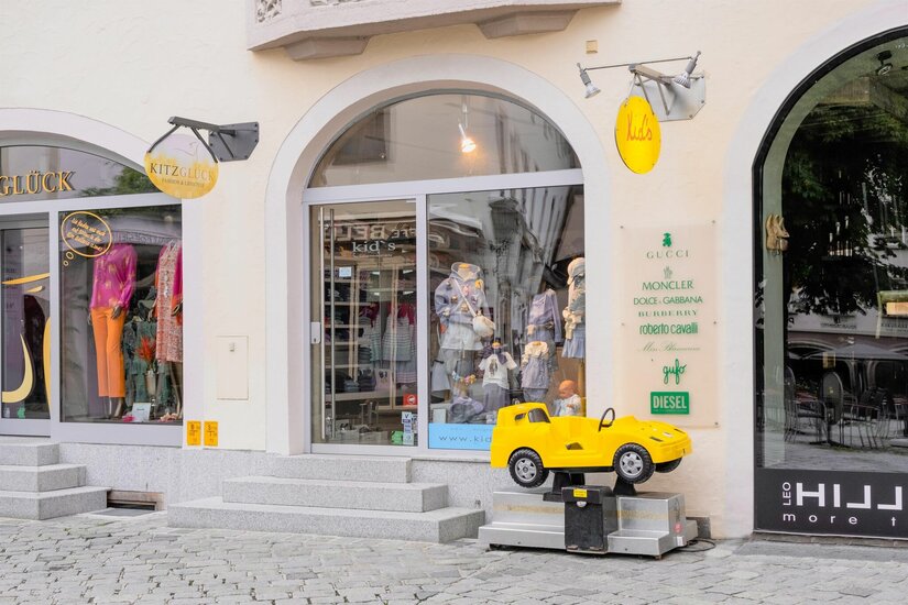Eingang eines Kinderbekleidungsgeschäfts mit Schaufensterpuppen und einem gelben Spielzeugauto davor.