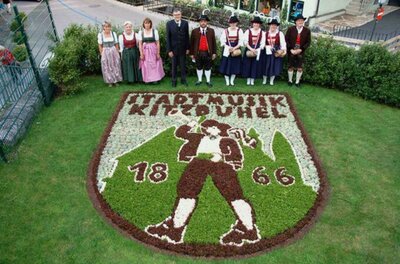 Jubiläumsgarten Eine Gruppe von Menschen in traditionellen Kleidern steht vor einem kunstvoll angelegten Blumenbeet in Form eines Wappens. Das Wappen zeigt einen Musiker und die Jahreszahlen 1866 und 2004.