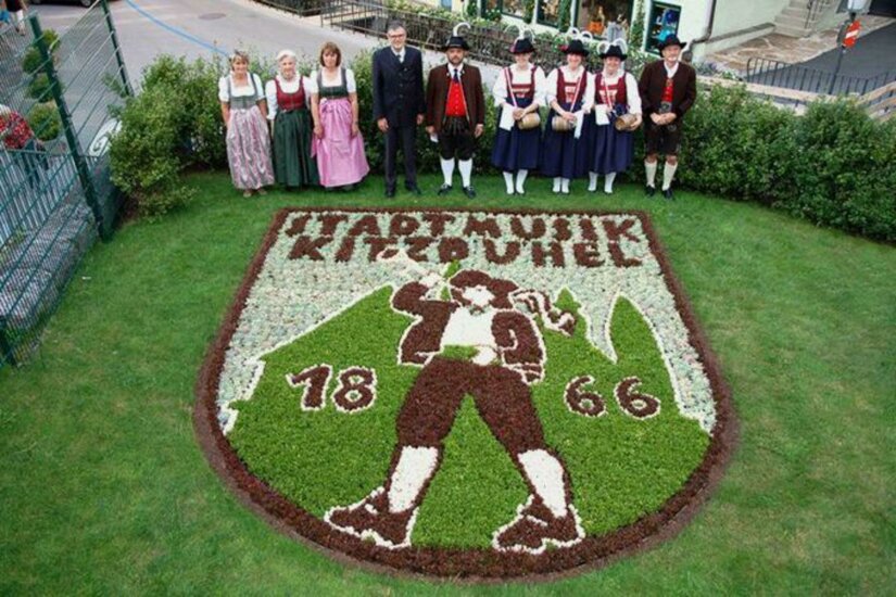 Jubiläumsgarten Eine Gruppe von Menschen in traditionellen Kleidern steht vor einem kunstvoll angelegten Blumenbeet in Form eines Wappens. Das Wappen zeigt einen Musiker und die Jahreszahlen 1866 und 2004.