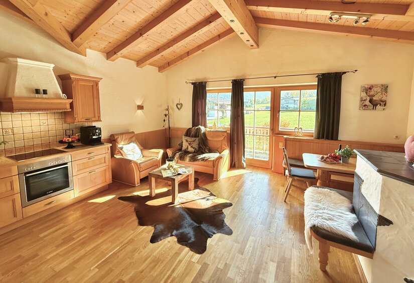 Ein gemütliches Wohnzimmer mit Holzdecke und einem großen Fenster. Die Einrichtung besteht aus bequemen Sesseln, einem Tisch und einer modernen Küche.
