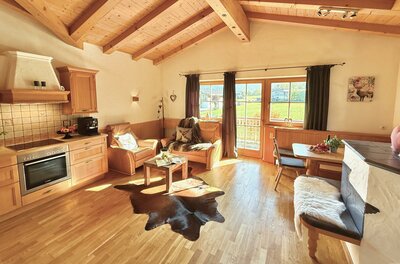 Ein gemütliches Wohnzimmer mit Holzdecke und einem großen Fenster. Die Einrichtung besteht aus bequemen Sesseln, einem Tisch und einer modernen Küche.