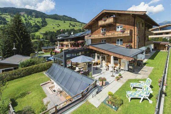 Alpines Holzhaus mit Sonnenterrasse, Liegestühlen und Blick auf grüne Berghänge.