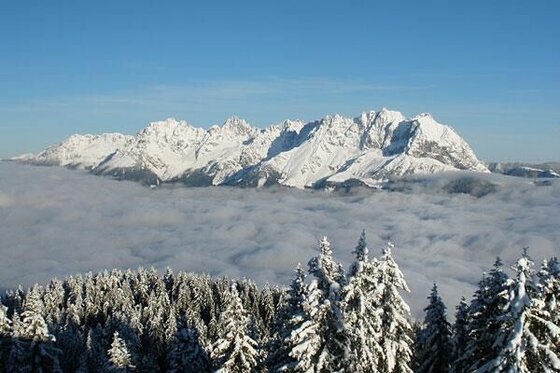 Winterpanorama am Noichl Jugendhotel Verschneite Berggipfel über einer dichten Wolkendecke mit schneebedeckten Tannen im Vordergrund.