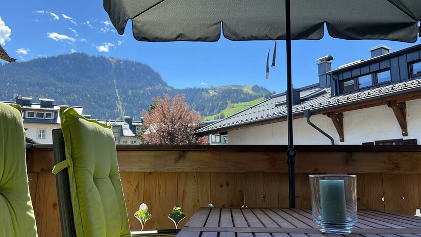 Balkonblick im Kitzalphouse Balkon mit Holztisch, grünem Stuhlpolster und Sonnenschirm vor bewaldeter Bergkulisse.