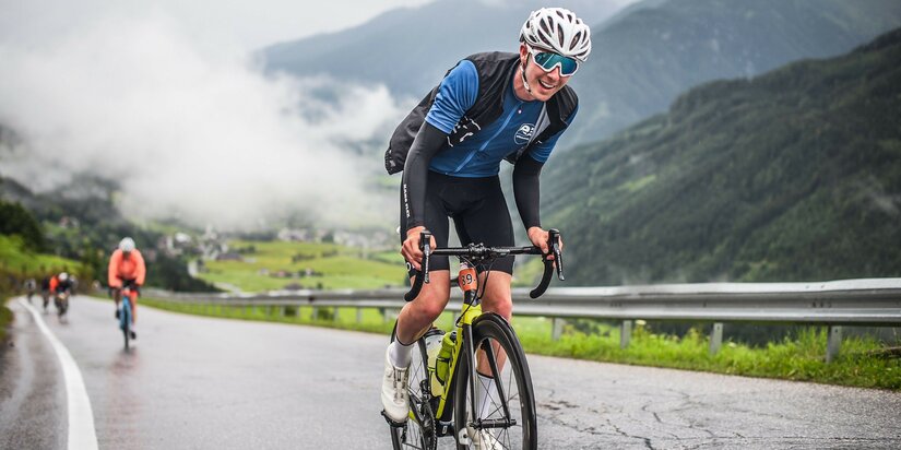 Radfahrer fährt lächelnd beim Kitzbüheler Radmarathon auf einer kurvenreichen Straße in Kitzbühel, umgeben von Bergen und Nebel.