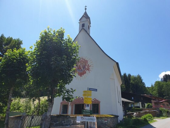 Weiße Kapelle mit Wandmalerei und gelben Wegweisern im Vordergrund unter blauem Himmel.