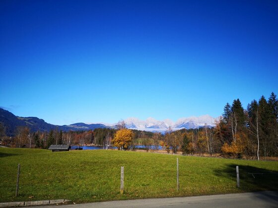 Blick auf den Schwarzsee Grüne Wiese mit Holzhütte vor einem See und schneebedeckten Bergen unter blauem Himmel.