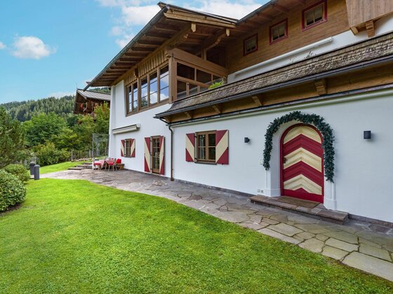 Alpines Wohnhaus mit weißer Fassade, rot-weiß gestreiften Fensterläden und einer verzierten Holztür vor einer Rasenfläche.