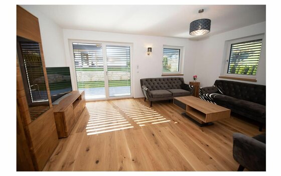 Wohnbereich Ferienhaus Reiter Modernes Wohnzimmer mit Parkettboden, zwei dunklen Sofas, Holzmöbeln und großen Fenstern mit Jalousien.