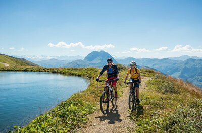 Zwei Mountainbiker fahren auf einem Schotterweg an einem Bergsee vorbei.