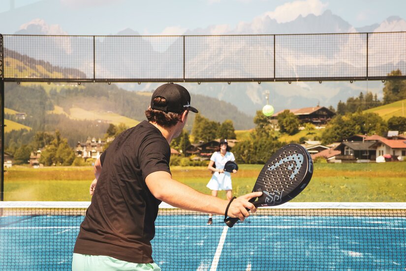 Padelplatz Eine Frau und ein Mann spielen Padeltennis auf dem blauen Platz. Zwischen ihnen ist das Netz, im Hintergrund sieht man Häuser und den Wilden Kaiser.