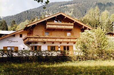 Zweistöckiges Alpenhaus aus Holz und weißem Putz mit Blumenbalkonen vor bewaldetem Berg.
