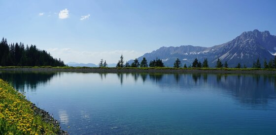 Ein klarer Bergsee mit Spiegelung der Bäume vor einem Gebirgsmassiv unter blauem Himmel.
