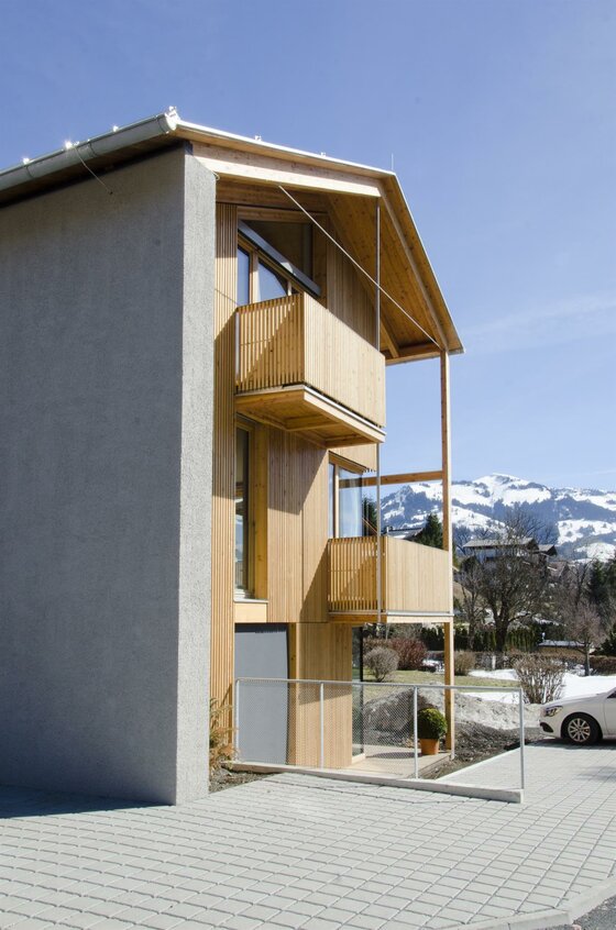 Kitzbühel Apartments Haus Wegener Modernes Gebäude mit Holzfassade, Balkonen und Blick auf verschneite Berge unter blauem Himmel.