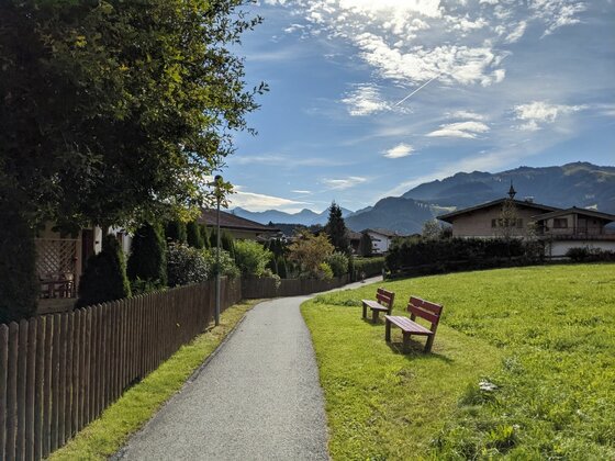 Wanderweg Rund um Reith Asphaltierter Spazierweg mit zwei Holzbänken neben einer Wiese und Wohnhäusern vor Gebirgskulisse.