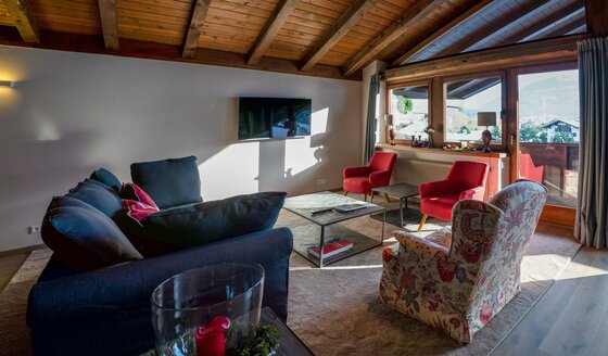 Wohnzimmer mit dunklem Sofa, roten Sesseln, Couchtisch und Fernseher unter einer Holzdecke.