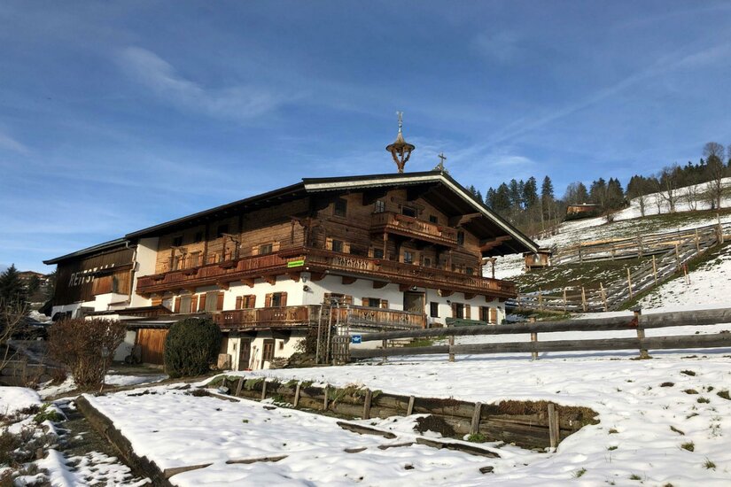 Henntalhof Kitzbühel im Winter Traditioneller Bauernhof aus Holz und weißem Putz in einer verschneiten Hanglage unter blauem Himmel.