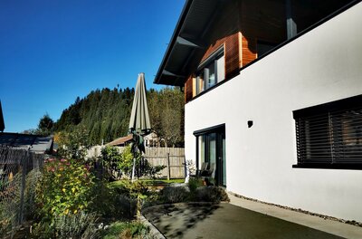 Modernes weißes Gebäude mit Holzfassade im Obergeschoss und Gartenbereich vor bewaldetem Hügel.