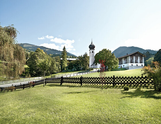 Kirche mit Zwiebelturm hinter einer Wiese mit Holzzaun vor bewaldeten Bergen.