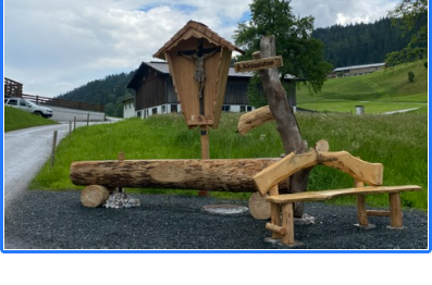 Ein Rastplatz mit Holzbank, Brunnen aus einem Baumstamm und einem Kruzifix vor einer Bergkulisse.