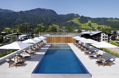 Ein langer Außenpool auf einer Dachterrasse mit Liegestühlen, Sonnenschirmen und Blick auf bewaldete Berge.