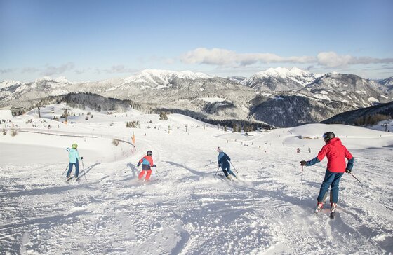 Eine Gruppe von Skifahrern fährt den schneebedeckten Hang hinunter. Im Hintergrund sind majestätische Berge und ein klarer Himmel zu sehen.