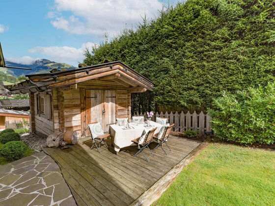 Gedeckter Esstisch auf einer Holzterrasse vor einer kleinen Blockhütte im Garten.