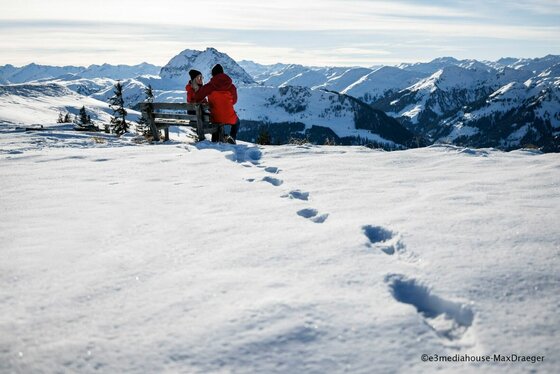 Winterwandern in Kitzbühel Zwei Personen sitzen auf einer Holzbank in einer verschneiten Berglandschaft mit Fußspuren im Vordergrund.