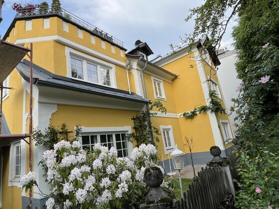 Schmiedhäusl Fassadenansicht Gelbes mehrstöckiges Gebäude mit weißen Fensterrahmen und blühenden Sträuchern im Vordergrund.