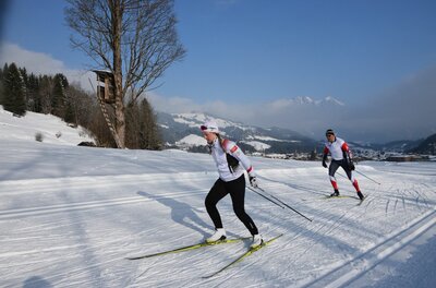 Zwei Personen beim Skilanglauf auf einer präparierten Loipe in einer verschneiten Berglandschaft.
