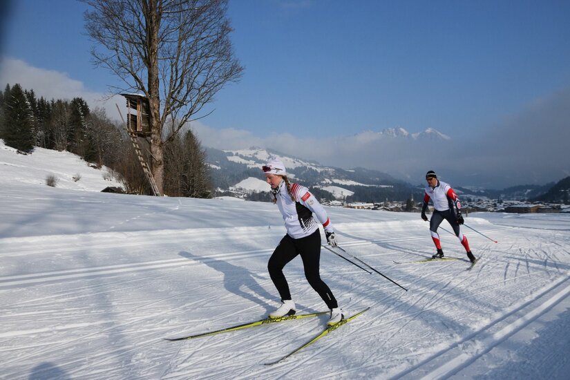Zwei Personen beim Skilanglauf auf einer präparierten Loipe in einer verschneiten Berglandschaft.