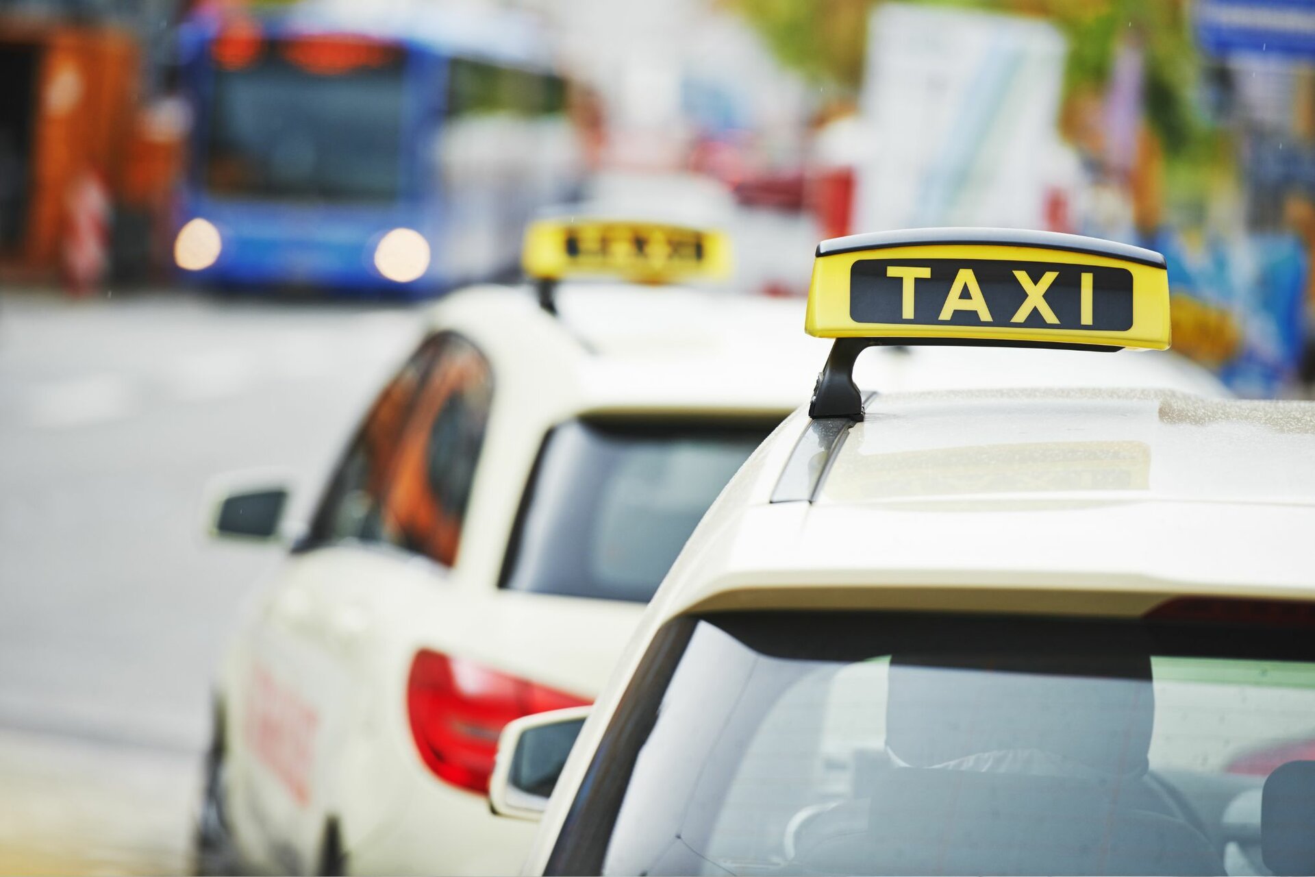 Zwei Taxis stehen an einer Straßenecke, während ein blauer Bus im Hintergrund vorbeifährt, was eine belebte Stadtatmosphäre schafft.