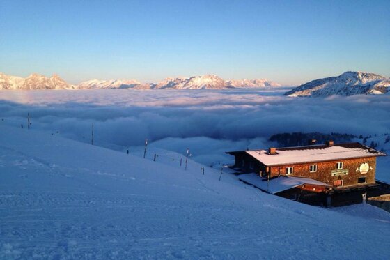Blick von einer verschneiten Skipiste auf die Hornköpflhütte über einer dichten Wolkendecke und Berggipfeln.