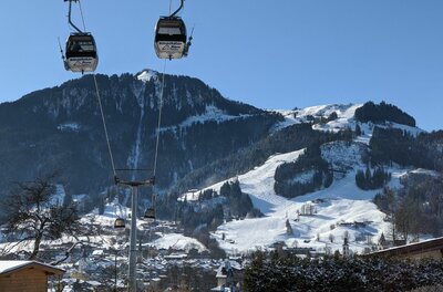 Zwei Gondeln einer Seilbahn schweben vor einem verschneiten Bergmassiv mit Skipisten unter blauem Himmel.