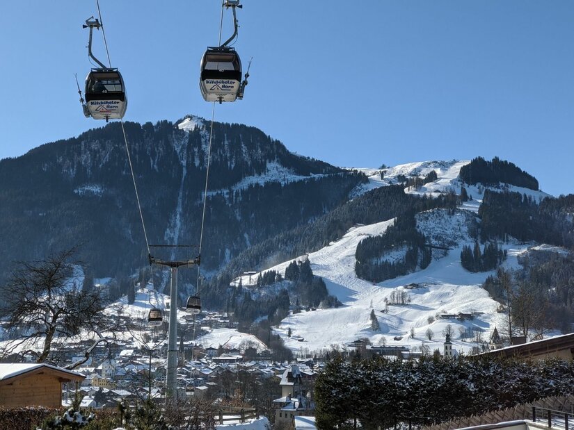 Zwei Gondeln einer Seilbahn schweben vor einem verschneiten Bergmassiv mit Skipisten unter blauem Himmel.