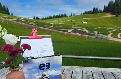 Holztisch mit Blumen und Speisekarte vor einem Bikepark im Hintergrund.