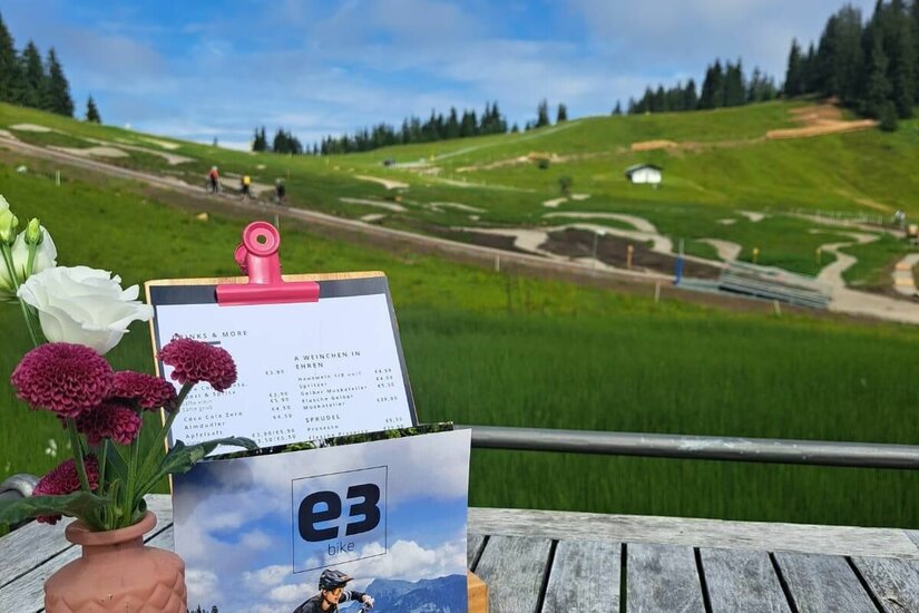Holztisch mit Blumen und Speisekarte vor einem Bikepark im Hintergrund.