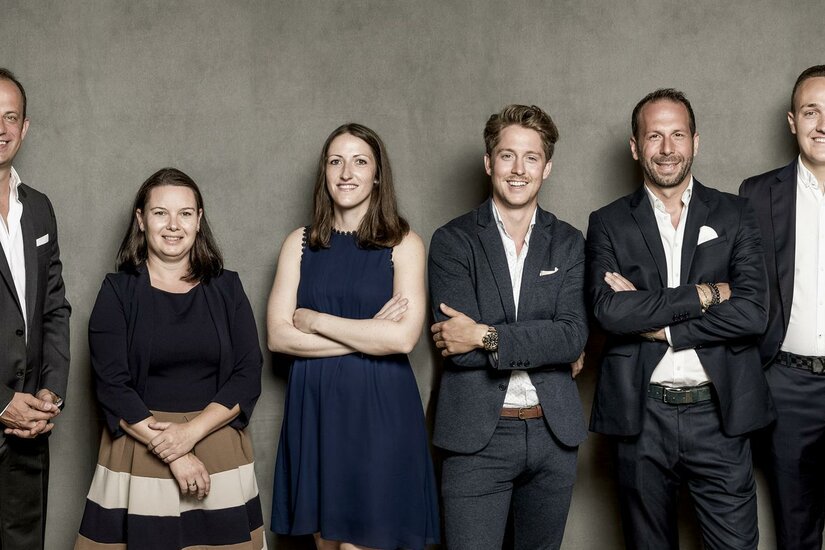 Cum Laude Immobilia Kitzbühel Eine Gruppe von sechs Personen steht vor einem grauen Hintergrund. Sie sind in formeller Kleidung gekleidet und lächeln.
