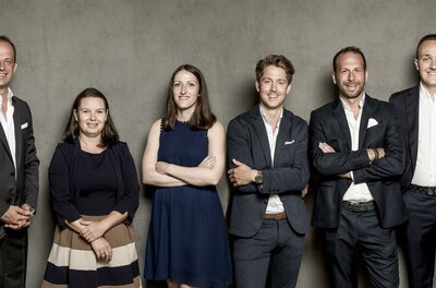 Eine Gruppe von sechs Personen steht vor einem grauen Hintergrund. Sie sind in formeller Kleidung gekleidet und lächeln.
