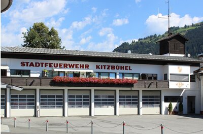Feuerwehr Ein Feuerwehrgebäude in Kitzbühel mit der Aufschrift "STADTFEUERWEHR KITZBÜHEL". Es gibt mehrere Fenster und einen klaren blauen Himmel im Hintergrund.