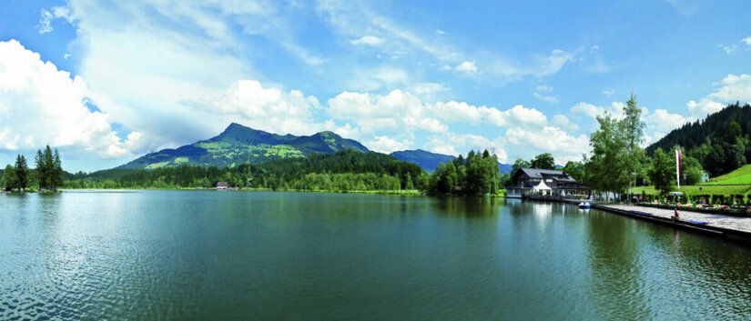 Schwarzsee im Sommer Panoramaaufnahme des Schwarzsees mit bewaldeten Bergen und einem Ufergebäude unter bewölktem Himmel.