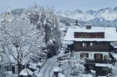 Verschneites Holzhaus in einem Alpendorf vor einer markanten, schneebedeckten Gebirgskette.